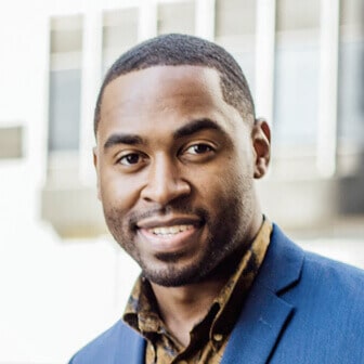 Professional headshot of a Black man with a beard, wearing a blue suit and a patterned shirt, smiling confidently outdoors.