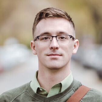 Headshot of a man with short brown hair and glasses, wearing a green collared shirt and sweater in an outdoor setting.