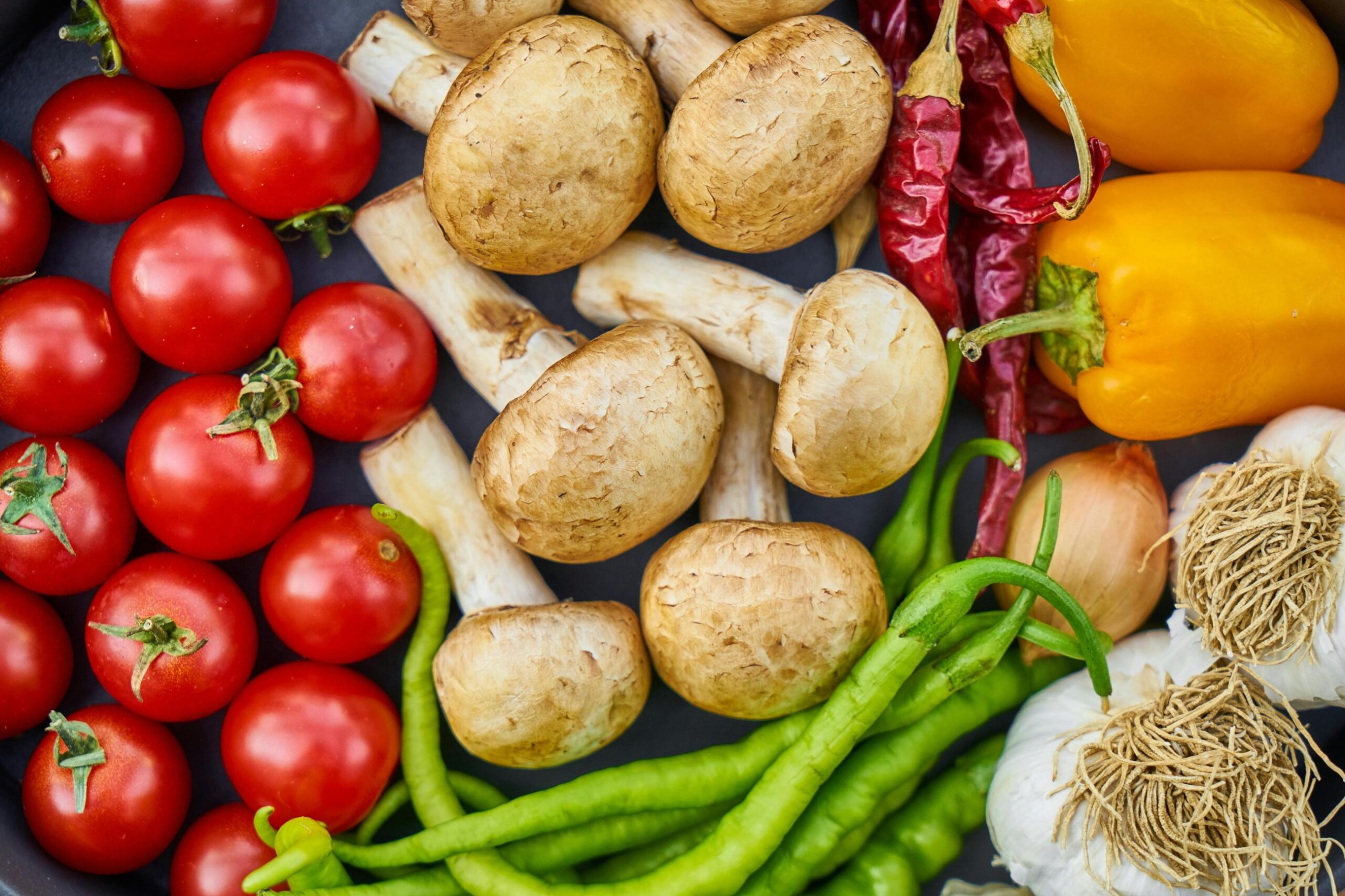 This is a vibrant overhead close-up of a variety of fresh produce, including bright red cherry tomatoes clustered on the left and large yellow bell peppers on the right.