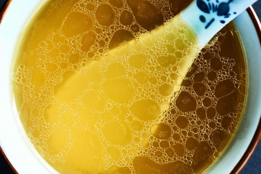 This is a high-angle, close-up photograph of a clear, golden-yellow liquid, likely soup or broth, filling a bowl with a white rim. The surface of the liquid is covered in a dense pattern of oil bubbles of varying sizes.