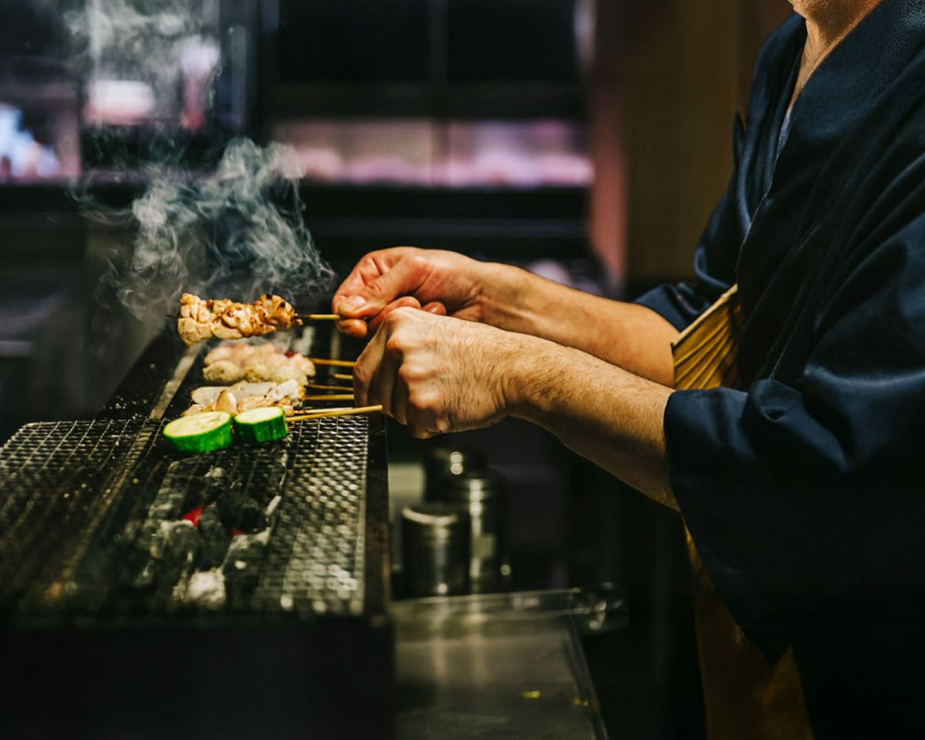 Close-up of a chef’s hand grilling chicken skewers over hot charcoal, with smoke rising and flames creating a warm, authentic izakaya cooking atmosphere.