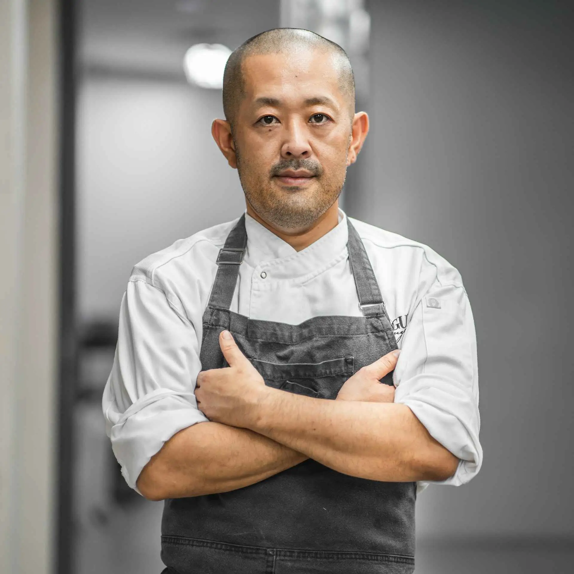 A half body photo of a Japanese chef with his arms crossed as he stands inside the kitchen of a restaurant.