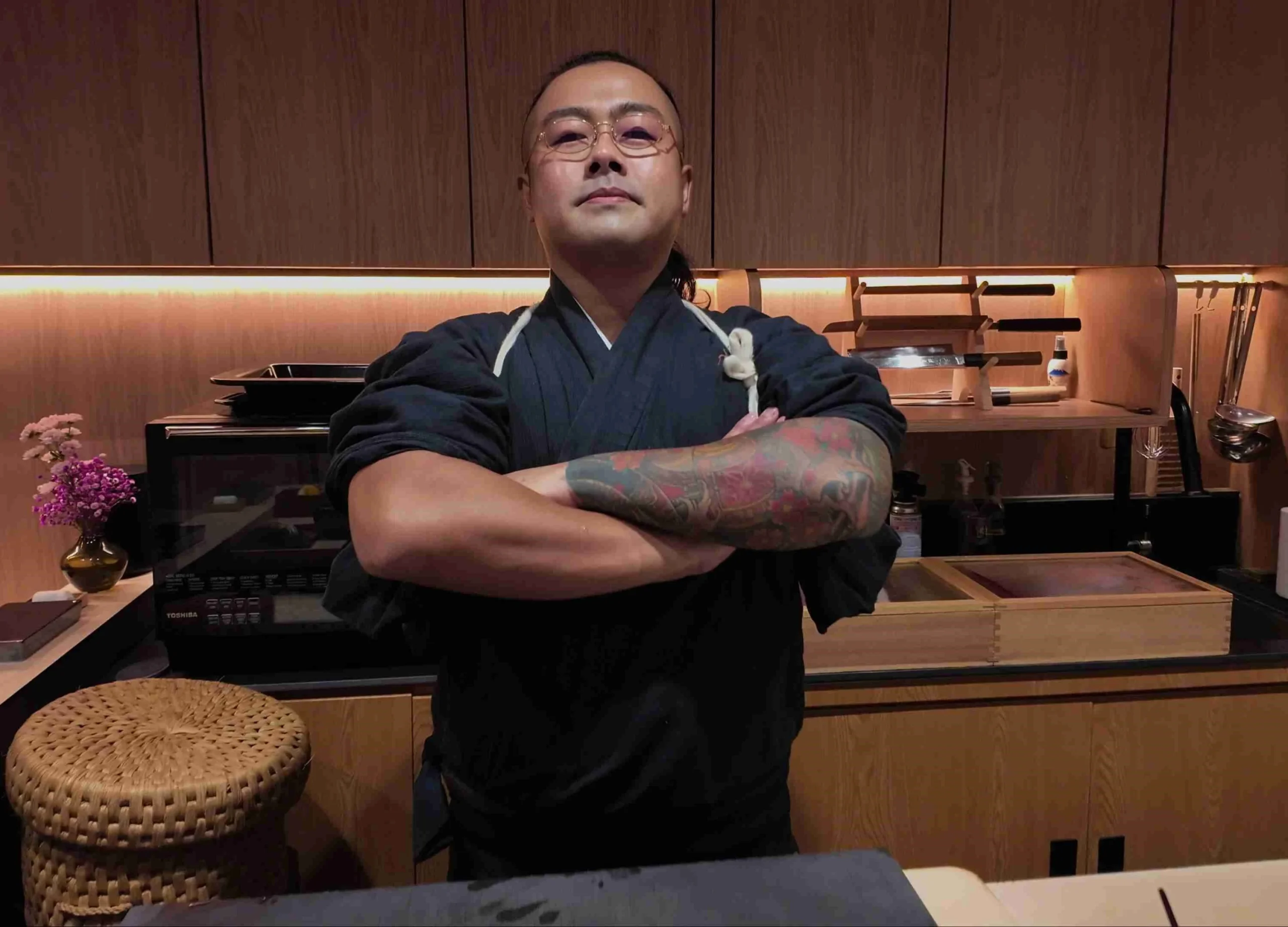 A professional portrait of Chef Masa standing with folded arms in a modern kitchen, featuring his distinctive arm tattoos and traditional dark chef attire.