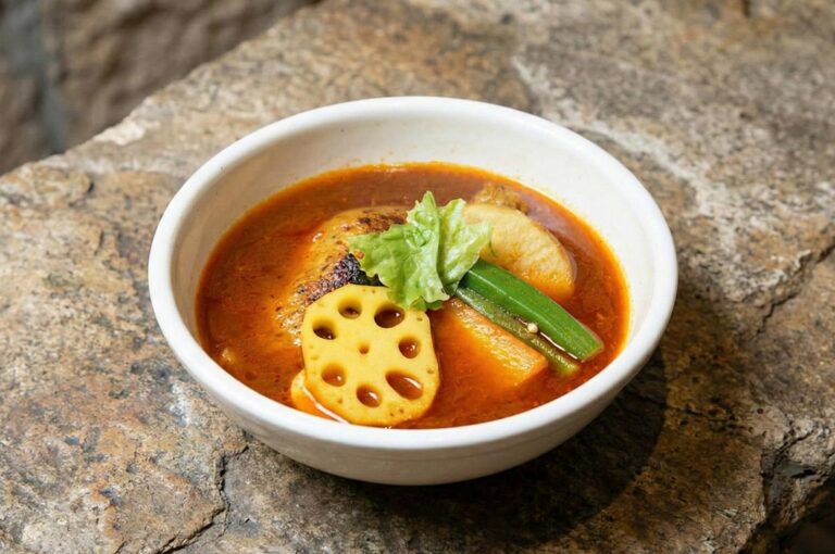 A bowl of Hokkaido-style soup curry with chicken, lotus root, okra, and carrots on a rustic stone background.