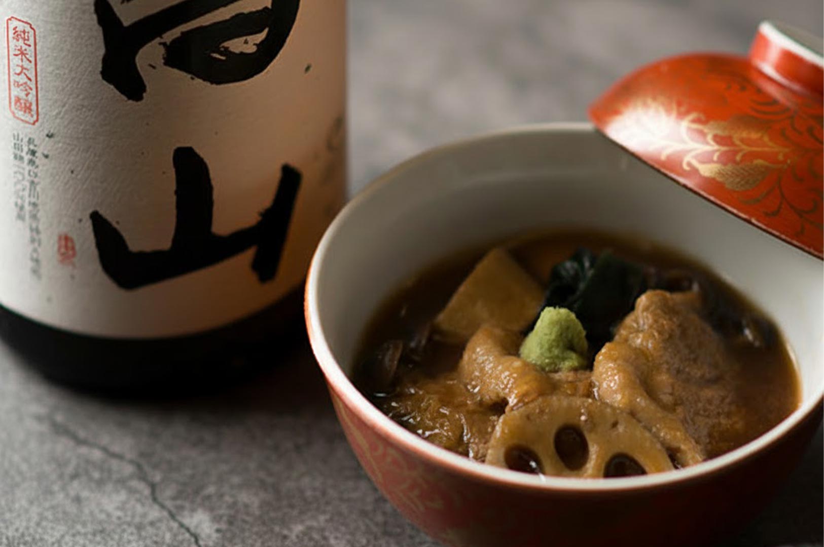 A ceramic bowl of Japanese soup curry containing lotus root and tofu topped with wasabi, sitting next to a traditional sake bottle.