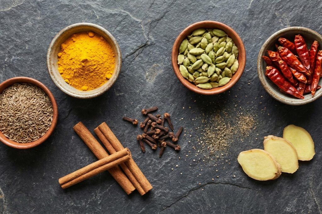 Flat lay of essential curry spices including turmeric powder, green cardamom, dried red chilies, cumin, cinnamon sticks, cloves, and fresh ginger.