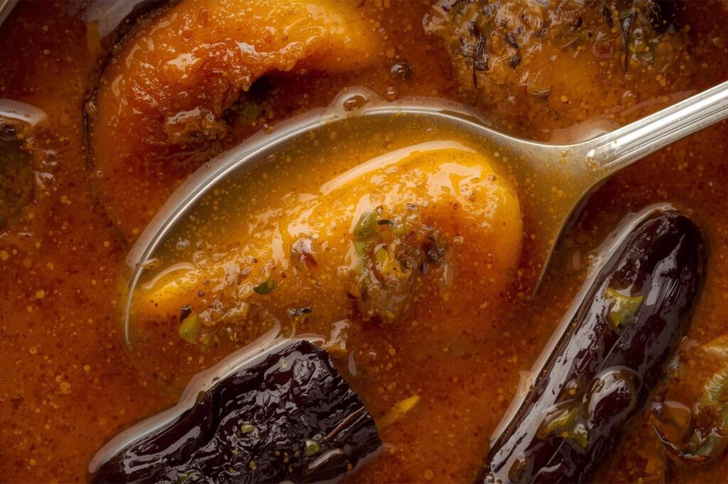 Extreme close-up of a silver spoon lifting a soft piece of pumpkin from a rich, oily curry broth containing dark dried chilies.