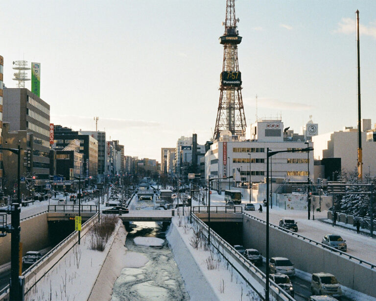 A cold winter morning in Sapporo with snow-covered streets and a frozen river leading toward the iconic Panasonic TV Tower under a clear blue sky.