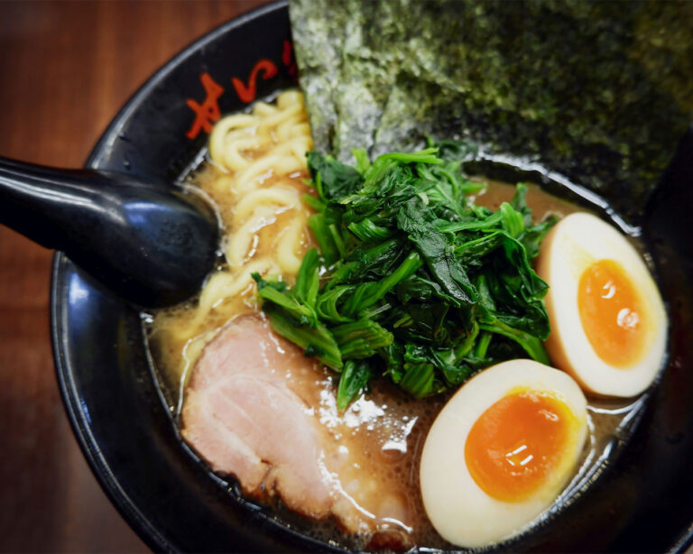 A bowl of rich Yokohama-style Iekei ramen with thick noodles, a slice of chashu pork, blanched spinach, a soft-boiled ajitama egg, and a large sheet of nori seaweed.