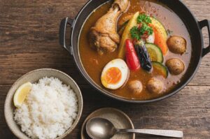 A bowl of Japanese soup curry with a chicken drumstick, soft-boiled egg, and roasted vegetables served with a side of white rice.