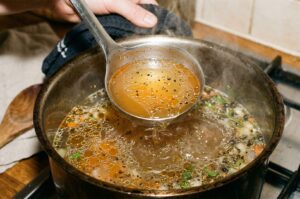 A clear, golden chicken broth with oil droplets and herbs being lifted by a metal ladle from a simmering pot on a stovetop.