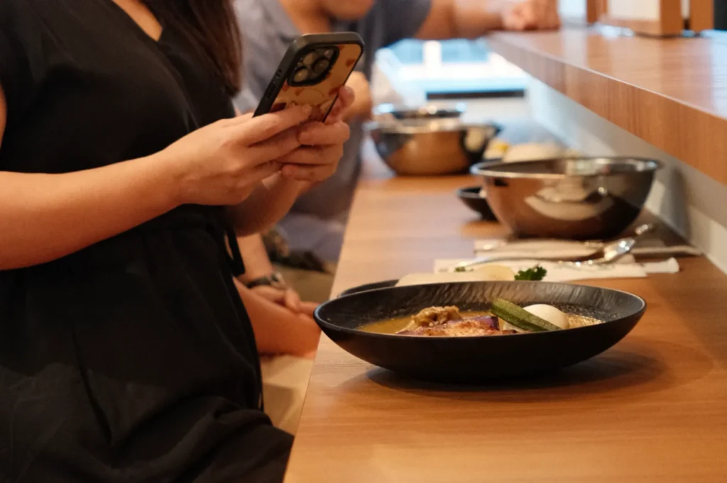 A person in a black dress is sitting at a table, holding a phone. In front of them is a bowl of food, and other people are visible in the background.
