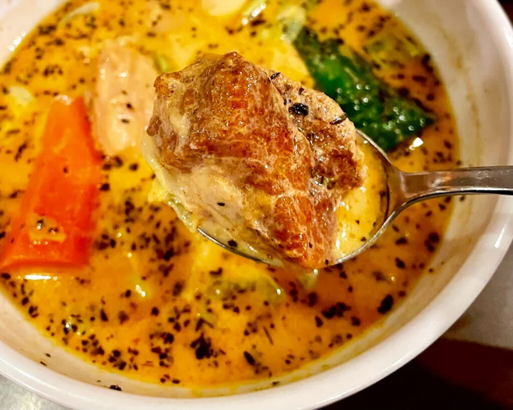 A spoon scooping rich, golden soup curry from a bowl, showing the thick texture of the broth with visible herbs and spices mixed inside.