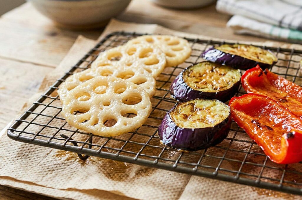 Slices of grilled lotus root, purple eggplant, and red bell peppers resting on a wire cooling rack over parchment paper.