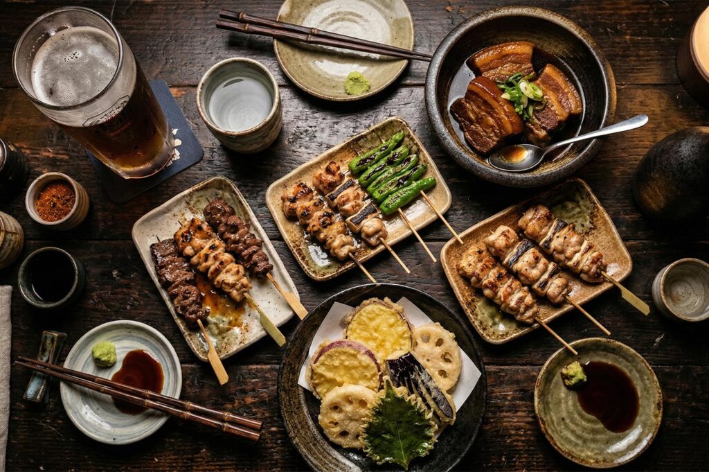 Top-down view of a wooden table filled with Japanese dishes including chicken yakitori skewers, vegetable tempura, braised pork belly, and a glass of beer.
