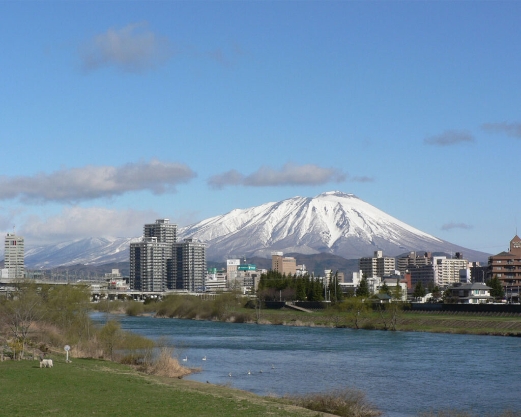 A scenic Japanese regional landscape with traditional buildings in the foreground and mountains in the background, under a clear sky, creating a calm and picturesque atmosphere.