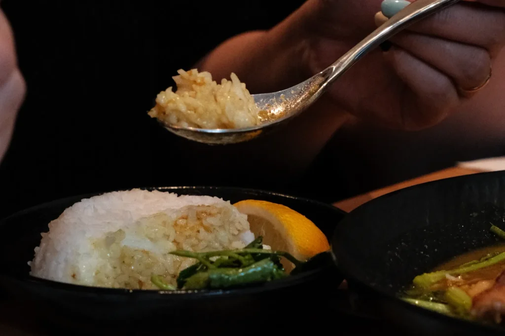 A person holding a spoon with a small serving of rice, next to a plate of rice with greens and a lemon wedge.