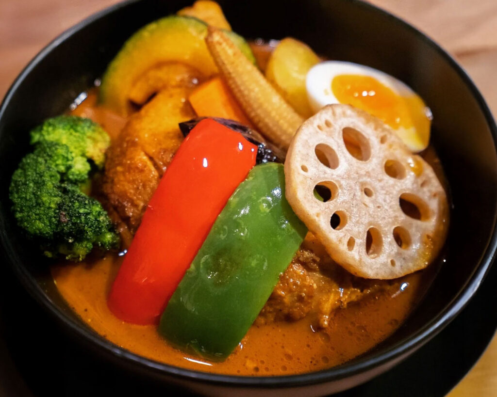 A bowl of Hokkaido soup curry filled with fresh seasonal vegetables, including broccoli, pumpkin, and carrots, served in a light, fragrant broth with rice on the side.