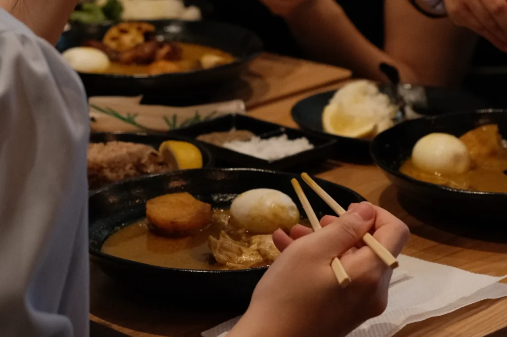 A close-up of hands holding chopsticks over a bowl of food, with several other dishes on the table, including a plate with a boiled egg and slices of lemon.