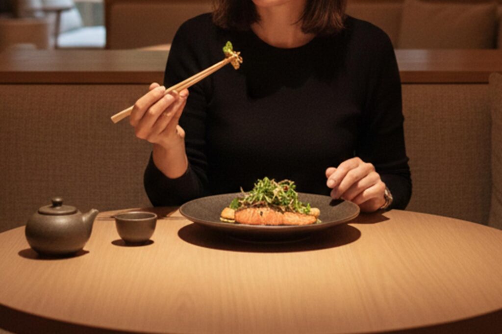A woman using chopsticks to eat a grilled salmon fillet topped with greens, served with a traditional clay teapot and cup.