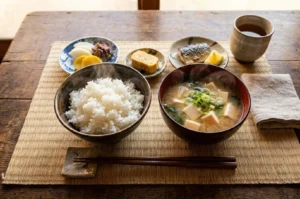 A traditional Japanese breakfast set featuring a steaming bowl of white rice, tofu miso soup, grilled mackerel, tamagoyaki, and pickles on a bamboo mat.
