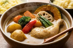 A steaming bowl of Japanese chicken soup curry featuring a whole chicken leg, roasted pumpkin, lotus root, okra, and broccoli.