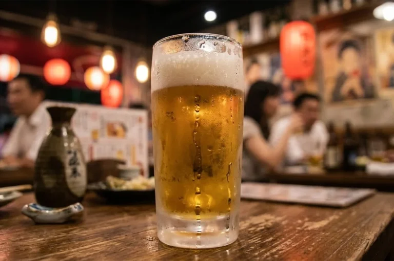 Close-up of a chilled Japanese lager in a frosted glass at a traditional yakitori restaurant with atmospheric indoor lighting and a sake carafe.