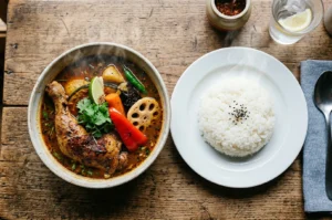 Overhead view of a bowl of Japanese soup curry with a roasted chicken leg, lotus root, and vegetables served with a side of white rice.