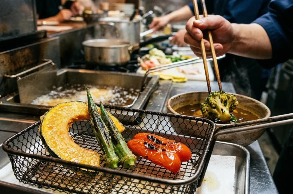 A chef using long wooden chopsticks to dip broccoli into sauce next to a basket of fried pumpkin, okra, and red bell peppers in a professional kitchen.