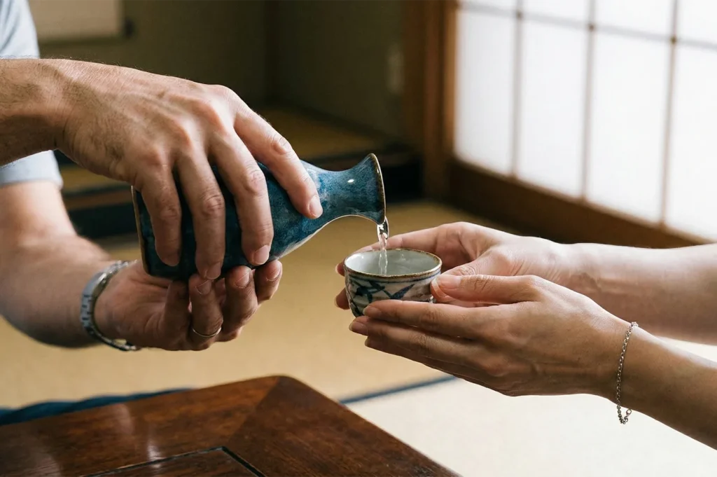 Close-up of a person pouring sake from a blue ceramic carafe into a small cup held by another person in a traditional Japanese room.