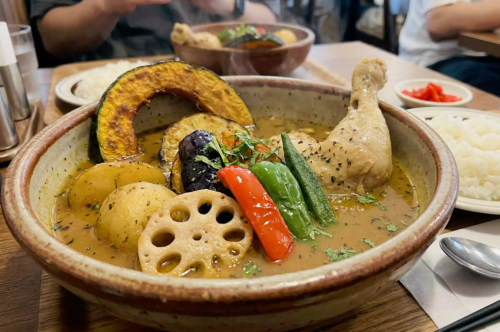 A detailed shot of a vibrant soup curry containing a chicken drumstick, fried lotus root, eggplant, and green peppers, with a bowl of rice in the background.