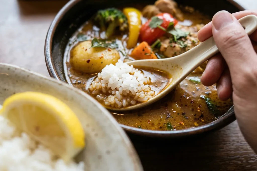 A close-up of a person using a ceramic spoon to scoop white rice into a bowl of rich Japanese soup curry filled with potatoes and colorful vegetables.