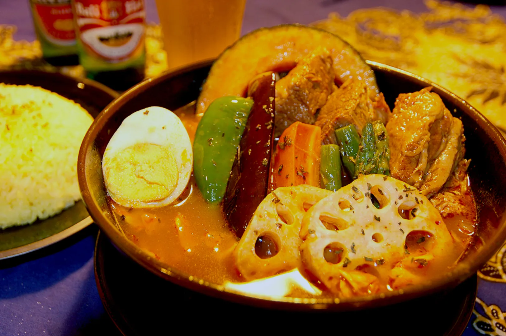 A colorful bowl of Japanese soup curry with boiled egg, lotus root, green pepper, and carrots. Yellow rice is on the side, creating a vibrant, appetizing scene.