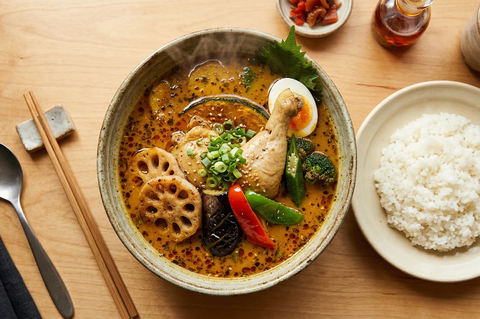 A top-down view of a bowl of Japanese Soup Curry featuring a chicken leg, lotus root, eggplant, and egg, served with a side of white rice