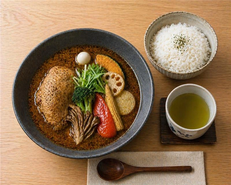 A bowl of Japanese Hokkaido soup curry with chicken leg, lotus root, and pumpkin, served with white rice and green tea.