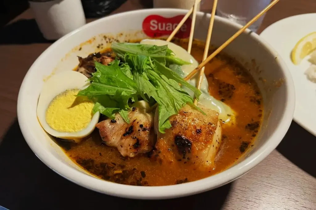 A bowl of rich, spicy broth with grilled chicken skewers, a halved boiled egg, greens, and noodles. The dish is vibrant and appetizing.