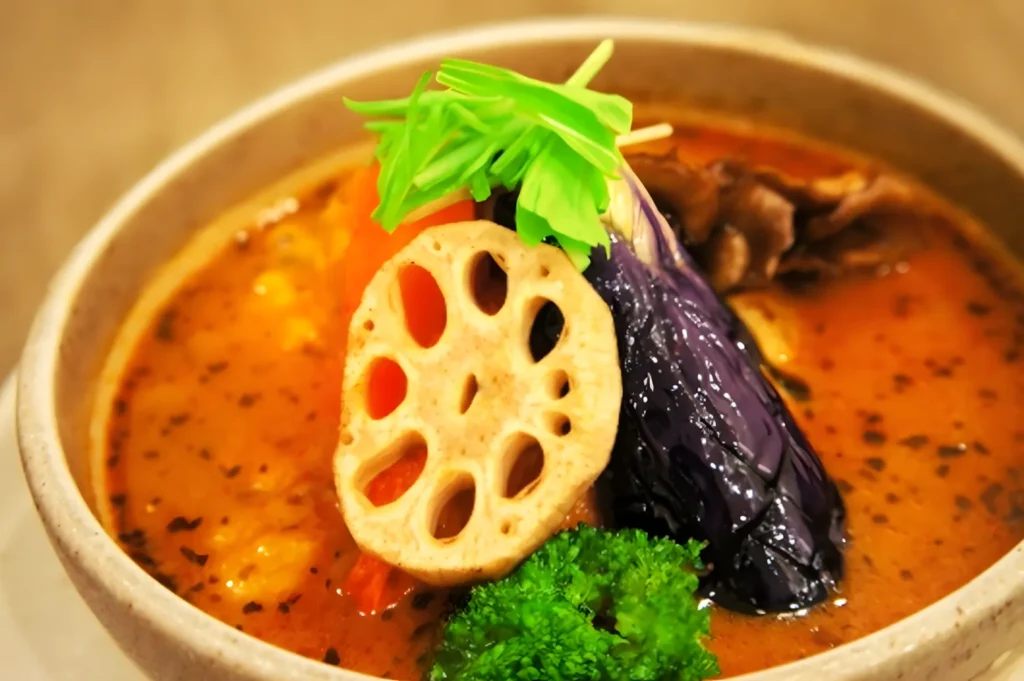 A colorful bowl of vegetable curry with vibrant orange broth, topped with sliced lotus root, purple eggplant, green broccoli, and fresh herbs.