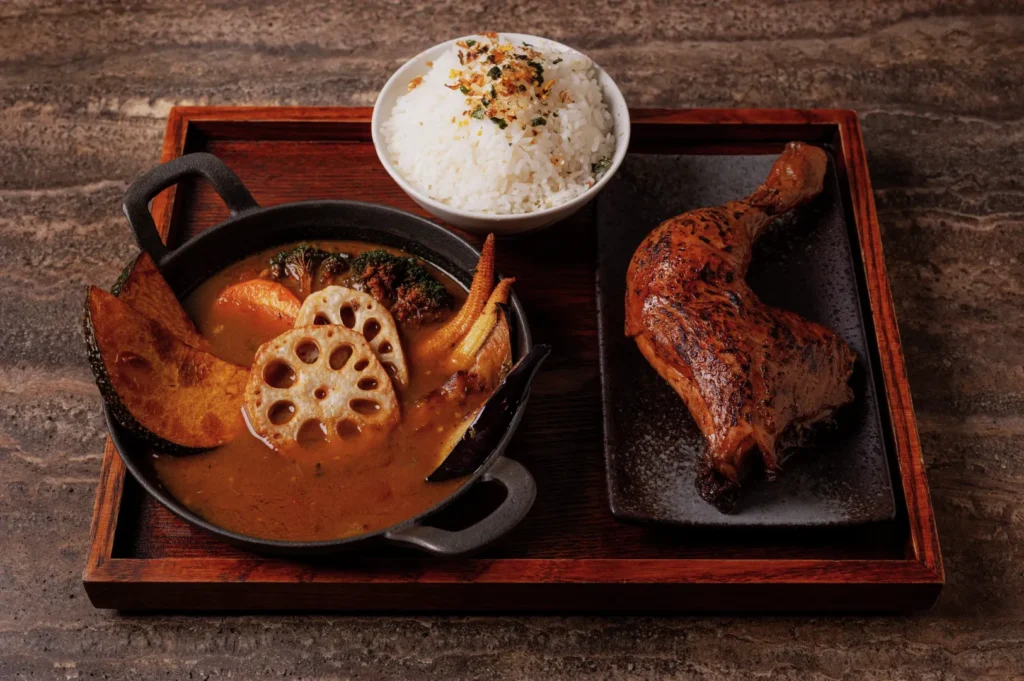 A wooden tray with spicy curry in a bowl filled with lotus root, vegetables, and chicken, a plate with roasted chicken leg, and a bowl of rice.
