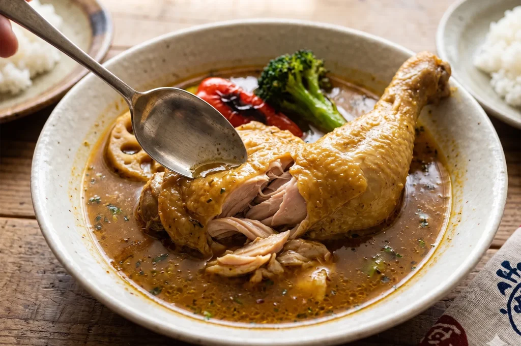 A spoon pulling apart a tender, fall-off-the-bone chicken leg in a bowl of aromatic Japanese soup curry.