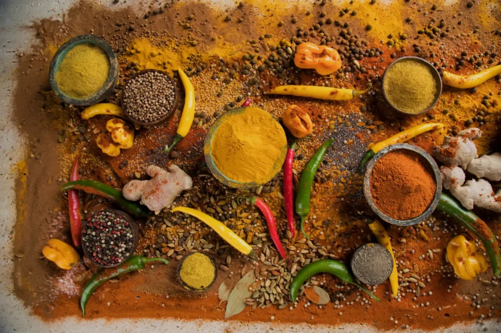 A vibrant mix of spices scattered on a surface, featuring bowls of turmeric and paprika, colorful chilies, ginger, and peppercorns, creating a rich and aromatic tapestry.