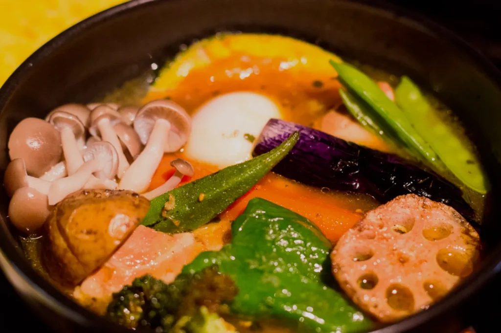 A colorful bowl of Japanese vegetable curry soup featuring shimeji mushrooms, sliced carrots, lotus root, egg, eggplant, and snow peas, creating a warm and hearty appearance.