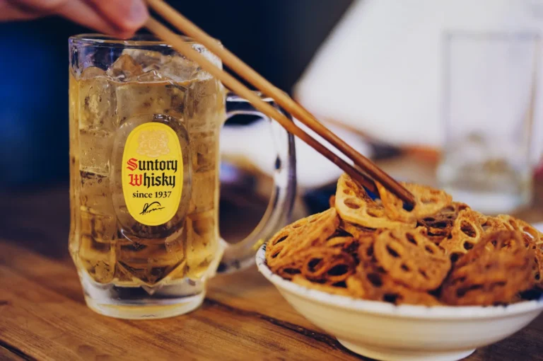A chilled Suntory Whisky mug with ice sits next to a bowl of fried lotus root chips. Chopsticks hover above, giving a casual, relaxed dining vibe.