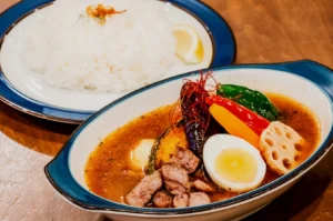 A vibrant bowl of soup curry with grilled chicken, boiled egg, and colorful vegetables, alongside a plate of white rice with a lemon wedge.