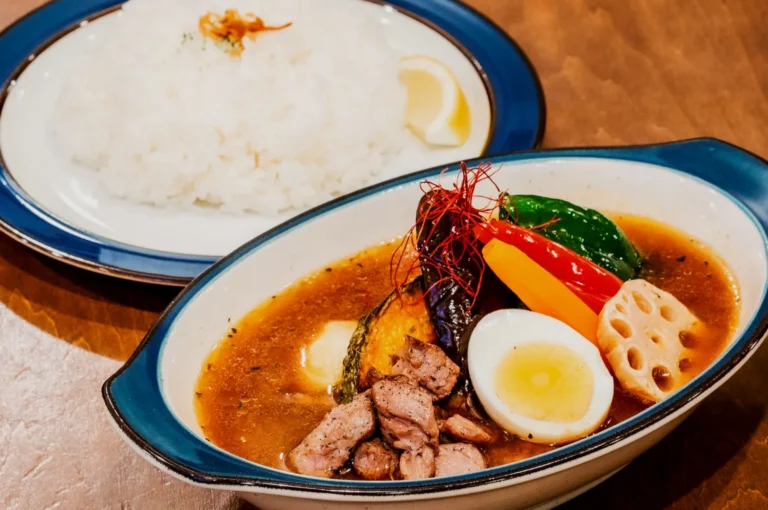 A vibrant bowl of soup curry with grilled chicken, boiled egg, and colorful vegetables, alongside a plate of white rice with a lemon wedge.