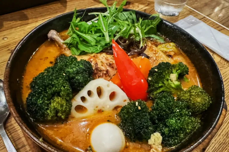A bowl of vibrant curry soup includes broccoli, red pepper, lotus root, leafy greens, and a poached egg in rich broth, set on a wooden table.
