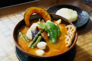 A vibrant bowl of vegetable curry soup with pumpkin, green peppers, lotus root, and beans, garnished with herbs. Served alongside a plate of rice.