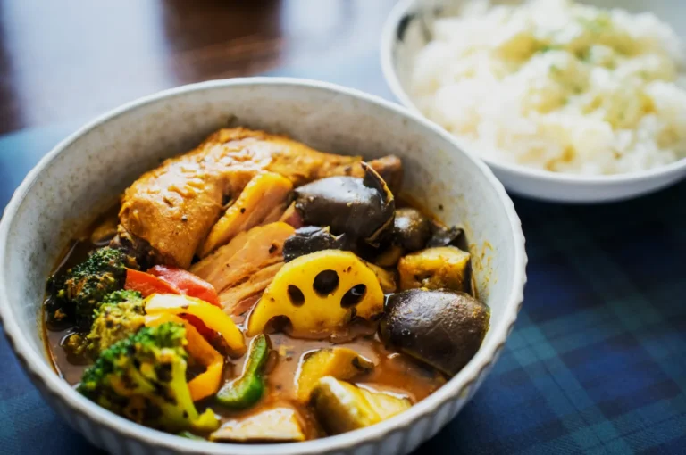 A bowl of vibrant curry soup with chunky vegetables, lotus root, a halved boiled egg, and diced green onions, evoking warmth and richness. Rice sits aside.