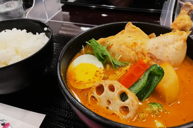 A vibrant bowl of red curry soup with a chicken leg, boiled egg, lotus root slice, vegetables, and herbs, accompanied by a bowl of white rice.