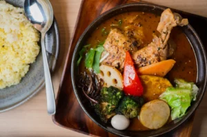 A vibrant bowl of Japanese curry soup with chicken leg, colorful vegetables, and quail egg. Served with a side of yellow rice and a spoon on a plate.