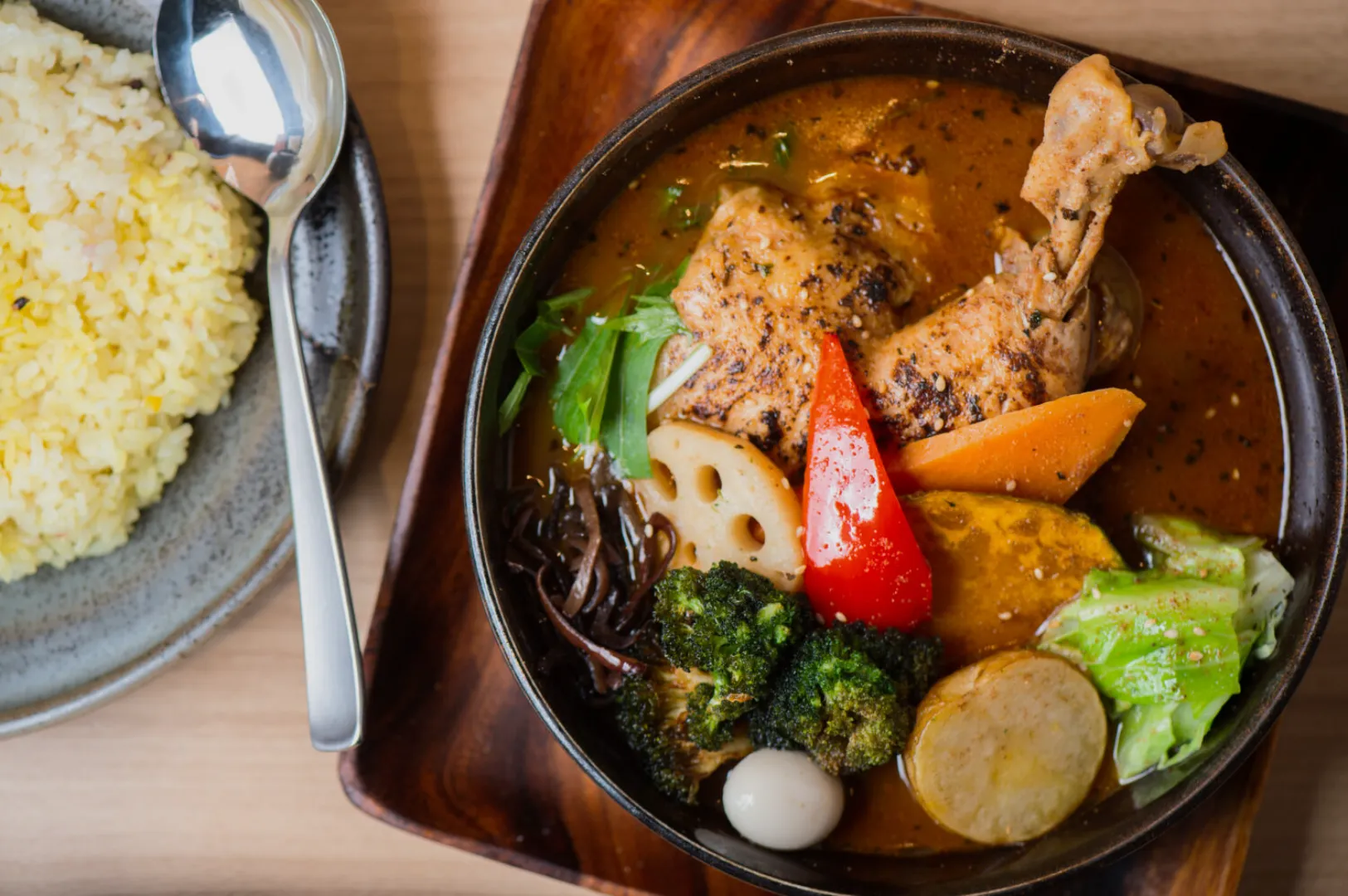 A vibrant bowl of Japanese curry soup with chicken leg, colorful vegetables, and quail egg. Served with a side of yellow rice and a spoon on a plate.